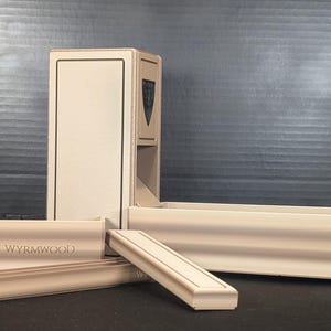 Puede incluir: Un conjunto de torre de dados y bandeja de color beige con un componente de torre vertical que presenta un emblema de escudo oscuro y una bandeja horizontal. La palabra WYRMWOOD está grabada en el frente de la bandeja. El set es de material claro.