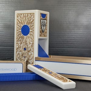 May include: A white and gold gaming dice tower and tray set featuring intricate carved patterns, a blue circular emblem, and a shield crest. The word WYRMWOOD is printed on a small rectangular white component. The set includes a tall tower, a long tray, and a separate blue grooved insert.