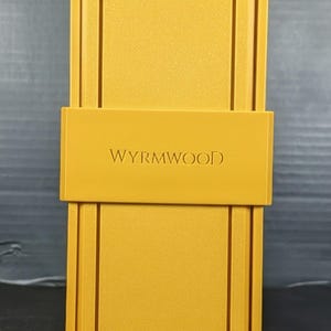 May include: A rectangular mustard yellow storage box with a sliding band across the center. The word WYRMWOOD is embossed in capital letters on the front of the band. The box features a clean, structured design with a matte finish and raised borders.