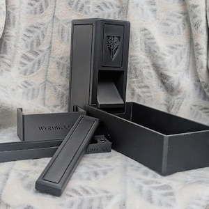 Puede incluir: Un conjunto de accesorios de juego en negro mate que incluye una torre de dados vertical con un emblema de árbol, una bandeja rectangular para dados y dos componentes separados con el texto Wyrmwood grabado en superficies de material oscuro.