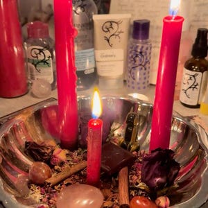 May include: A silver-toned scalloped bowl containing three lit red taper candles, dried rose petals, cinnamon sticks, smooth polished stones, and a small dark chocolate square. The arrangement is set against a background of various skincare bottles.