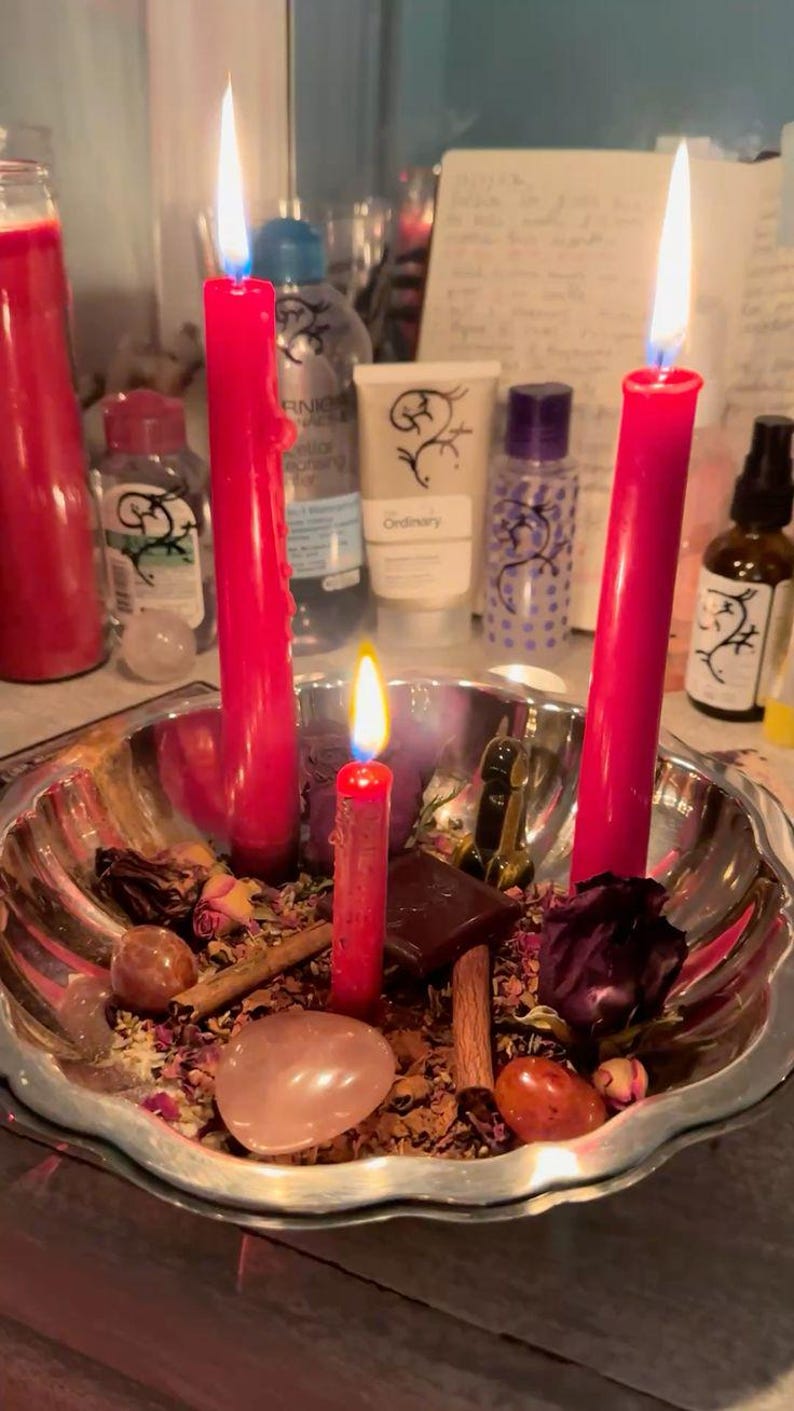 May include: A silver-toned scalloped bowl containing three lit red taper candles, dried rose petals, cinnamon sticks, smooth polished stones, and a small dark chocolate square. The arrangement is set against a background of various skincare bottles.