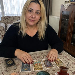 May include: A person with blonde hair wearing a black long-sleeved shirt sits at a table with four tarot cards laid out, a deck of cards, a small lit candle, three polished stones, a small ceramic bowl, and a glass of tea on a saucer.