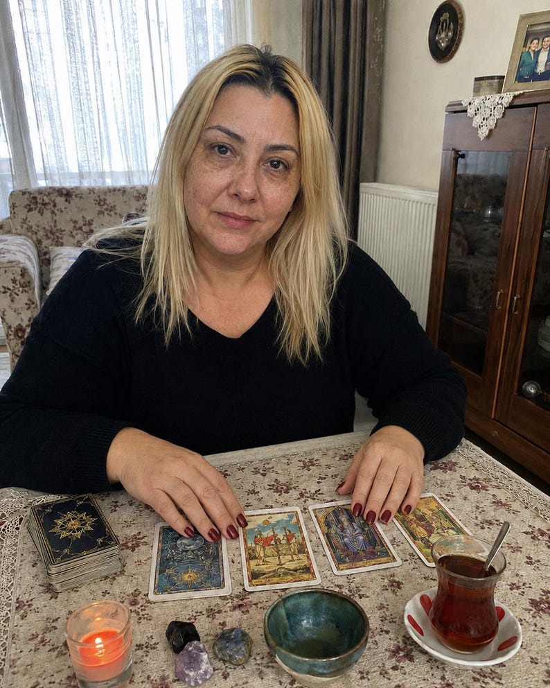 May include: A person with blonde hair wearing a black long-sleeved shirt sits at a table with four tarot cards laid out, a deck of cards, a small lit candle, three polished stones, a small ceramic bowl, and a glass of tea on a saucer.
