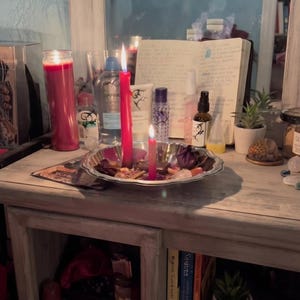 May include: A silver-toned metal tray holds two lit red candles, dried flower petals, and small crystals. Behind the tray are various skincare bottles, a small potted succulent, and an open notebook with handwritten text, all reflected in a mirror.