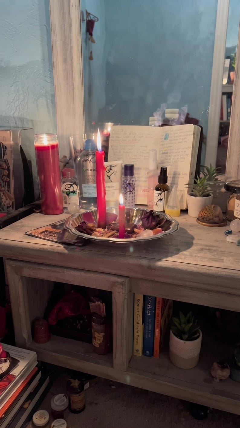 May include: A silver-toned metal tray holds two lit red candles, dried flower petals, and small crystals. Behind the tray are various skincare bottles, a small potted succulent, and an open notebook with handwritten text, all reflected in a mirror.