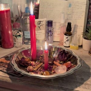 May include: A silver-toned scalloped metal bowl containing two lit red candles, dried flower petals, cinnamon sticks, and various polished stones. The bowl sits on a surface surrounded by skincare bottles, a notebook, and other household items.