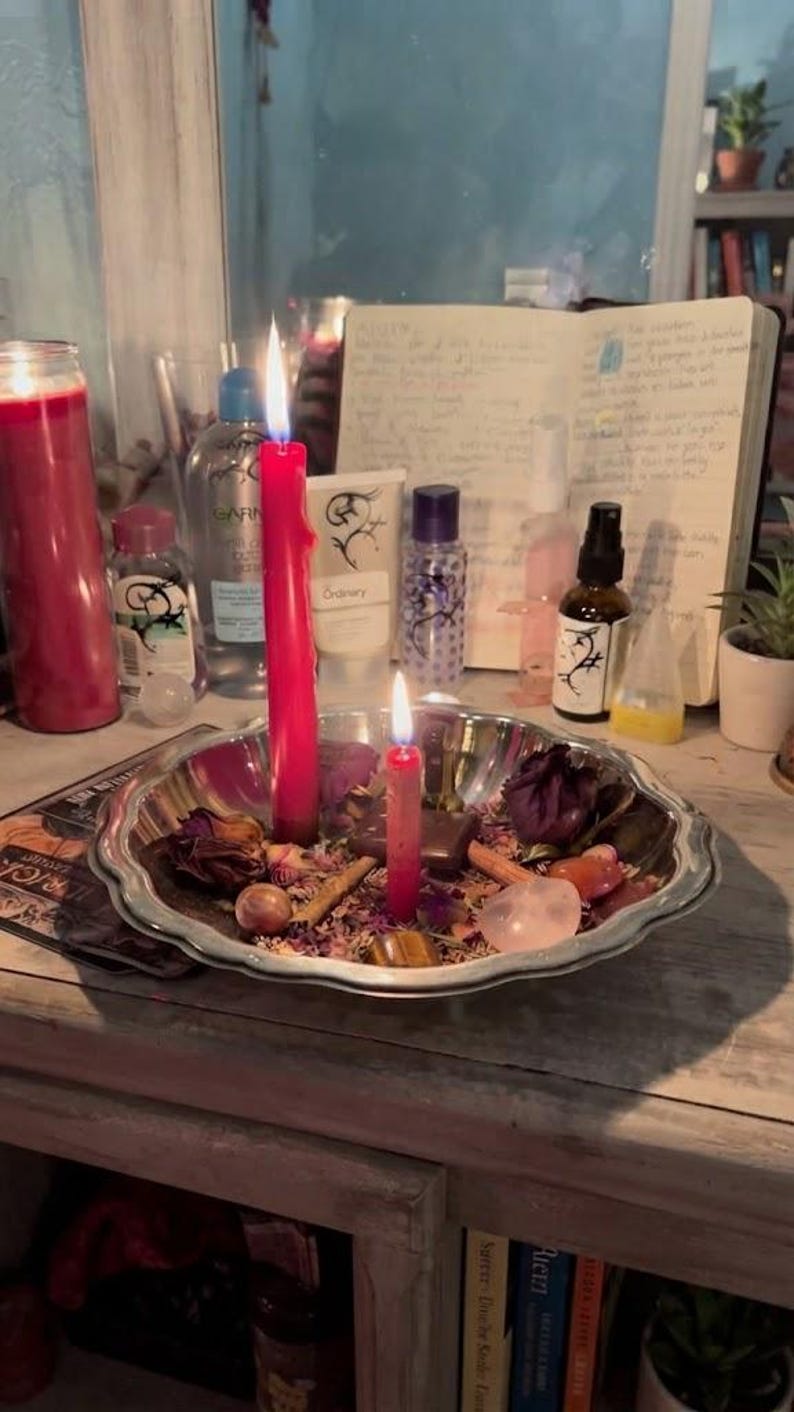 May include: A silver-toned scalloped metal bowl containing two lit red candles, dried flower petals, cinnamon sticks, and various polished stones. The bowl sits on a surface surrounded by skincare bottles, a notebook, and other household items.