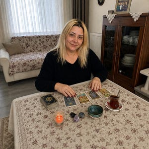 May include: A person with blonde hair wearing a black long-sleeved shirt sits at a table covered with a floral tablecloth. On the table are four tarot cards, a deck of cards, a small lit orange candle, three polished stones, and a glass of tea.