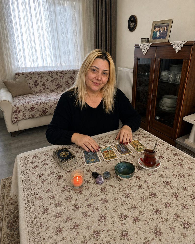 May include: A person with blonde hair wearing a black long-sleeved shirt sits at a table covered with a floral tablecloth. On the table are four tarot cards, a deck of cards, a small lit orange candle, three polished stones, and a glass of tea.