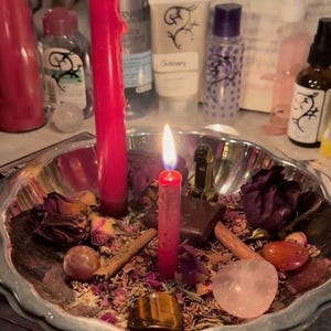 May include: A silver-toned metal bowl filled with dried flower petals, cinnamon sticks, polished gemstones, and a small dark square object. Two red candles are lit and standing upright within the bowl, surrounded by the scattered botanical items.
