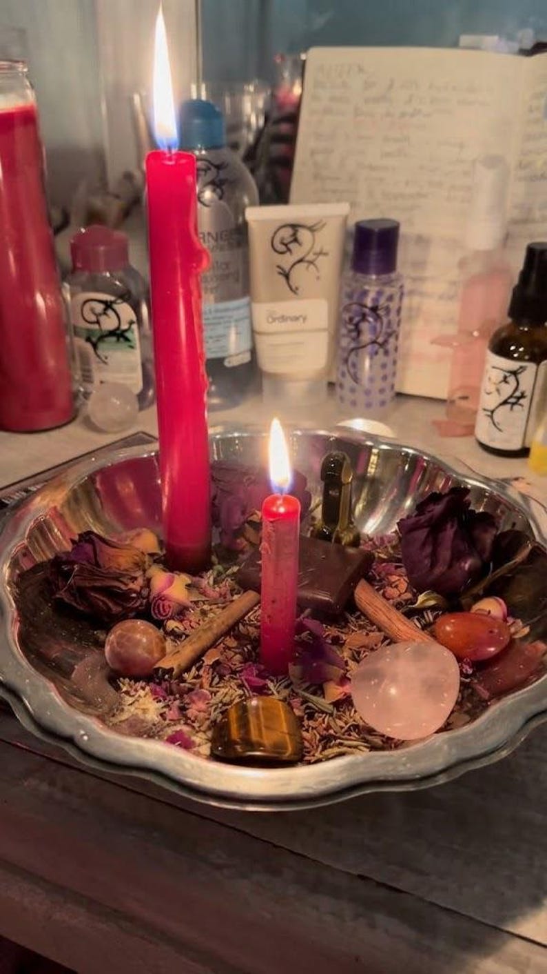 May include: A silver-toned metal bowl filled with dried flower petals, cinnamon sticks, polished gemstones, and a small dark square object. Two red candles are lit and standing upright within the bowl, surrounded by the scattered botanical items.