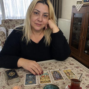 May include: A set of tarot cards laid out on a floral tablecloth, including a deck, four individual cards, a small lit candle, three polished stones, a small ceramic bowl, and a glass of dark tea on a saucer with red accents.