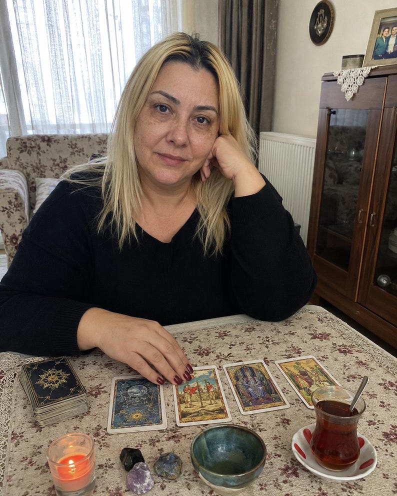 May include: A set of tarot cards laid out on a floral tablecloth, including a deck, four individual cards, a small lit candle, three polished stones, a small ceramic bowl, and a glass of dark tea on a saucer with red accents.