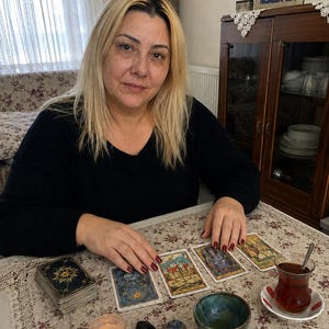 May include: A person with blonde hair wearing a black long-sleeved shirt sits at a table with a deck of tarot cards, three laid-out cards, a small lit candle in a glass holder, three small polished stones, a ceramic bowl, and a glass of tea.