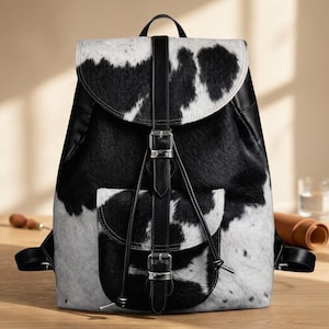 May include: A backpack featuring a black and white cowhide pattern with a textured fur-like appearance. The bag includes a top flap, a front pocket, and vertical black leather straps with silver-toned metal buckles and drawstring closures.