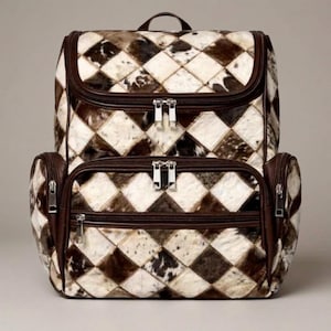 May include: A backpack featuring a diamond-patterned exterior made of brown, white, and black cowhide patches. The bag includes dark brown leather trim, multiple zippered compartments with silver-toned hardware, and a top handle for carrying.