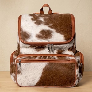 May include: A backpack featuring a brown and white cowhide pattern with a textured fur-like finish. The bag includes a top handle, a large main compartment with a zipper closure, and a smaller front pocket, all accented with brown leather trim.