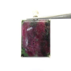 May include: A rectangular ruby zoisite pendant featuring a polished surface with deep magenta, dark green, and black mottled inclusions. The stone is held by a silver-toned metal bail at the top, showcasing a smooth, flat, and uniform shape.