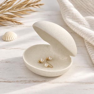 May include: A cream-colored, speckled ceramic trinket dish shaped like an open clam shell. The dish features a smooth, matte finish and contains a pair of gold-toned earrings with small white pearls resting inside the bottom shell section.