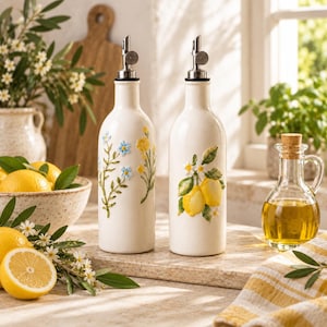 Op de afbeelding: Twee witte keramische oliedispensers met metalen schenktuiten. Eén fles heeft een reliëf bloemendesign met blauwe en gele bloemen, terwijl de andere een reliëfmotief van citroenen en groene bladeren toont. Beide flessen hebben een cilindrische vorm.