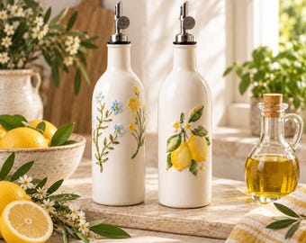 May include: Two white ceramic oil dispensers with metal pour spouts. One bottle features a raised floral design with blue and yellow flowers, while the other displays a raised lemon and green leaf motif. Both bottles have a smooth, cylindrical body.