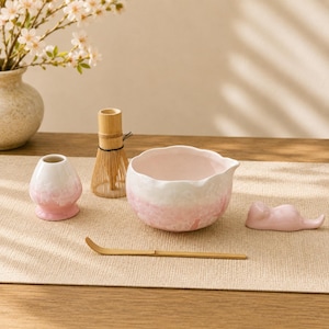 Op de afbeelding: Een matcha-theeset bestaande uit een lichtroze en witte keramische kom met een geschulpte rand, een bijpassende keramische gardehouder, een bamboe garde, een bamboe theeschepje en een kleine roze keramische dierenfiguur als lepelsteun.