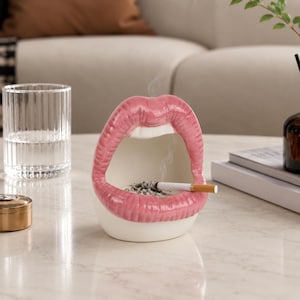 Puede incluir: Un cenicero de cerámica con forma de boca abierta, labios de color rosa brillante y dientes blancos. El interior de la boca sirve como recipiente, conteniendo ceniza gris y un cigarrillo encendido con filtro blanco y cuerpo beige.