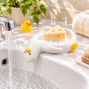 May include: A white ceramic soap dish shaped like a duck with a yellow beak and feet. The dish holds a wedge of yellow soap textured to resemble a piece of Swiss cheese with holes. The duck head extends over the edge of a white sink basin.