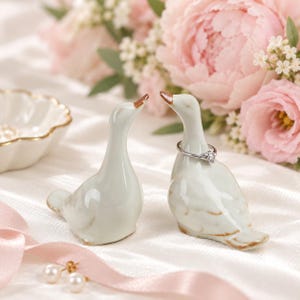 May include: Two white ceramic duck figurines with brown-tipped beaks and tails. One duck wears a silver ring with a small clear stone around its neck. The smooth, glossy surface of the birds reflects light, and they are positioned side by side.