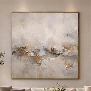 Puede incluir: Una gran pintura abstracta cuadrada con capas texturizadas en tonos crema, beige, gris y oro metálico, enmarcada con un borde dorado fino, colgada en una pared neutra sobre un largo sofá seccional color crema y una mesa de centro de mármol.