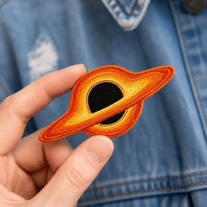May include: An embroidered patch featuring a black hole with a glowing orange and yellow accretion disk. The design depicts a dark circular center surrounded by concentric rings of vibrant warm-toned thread, creating a detailed space-themed motif.