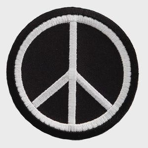 May include: A circular embroidered patch featuring a black background with a white stitched border and a white peace symbol centered within the design. The embroidery is dense and uniform, creating a clean contrast between the black and white.