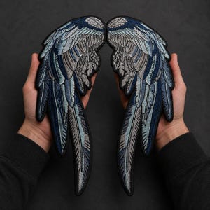 以下が含まれることがあります： 鳥の翼の形をした、精巧な刺繍ワッペンの一対。翼にはネイビーブルー、ライトブルー、グレー、ホワイトの色合いで複雑なステッチが施されており、黒い縁取りが羽の模様と翼全体の形状を際立たせています。