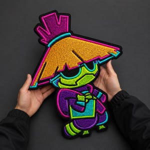 May include: A large embroidered patch featuring a stylized character wearing a wide, golden-yellow conical hat with a bright pink top accent. The character has a lime green face, a purple outfit with cyan trim, and a black border outline.