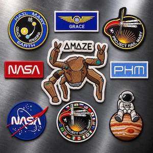 以下が含まれることがあります： 宇宙をテーマにした刺繍ワッペンのコレクション。AMAZEの文字が入った茶色のカニのような生き物、NASAのロゴ、プロジェクト・ヘイル・メアリーのミッションエンブレム、翼のある青いGraceワッペン、惑星に座る宇宙飛行士などが含まれています。