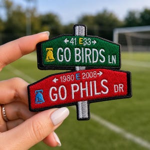 May include: A two-tiered embroidered patch shaped like street signs. The top green sign reads 41 E33 GO BIRDS LN with a yellow bell icon. The bottom red sign reads 1980 E 2008 GO PHILS DR with a blue bell icon, both mounted on a gray post.