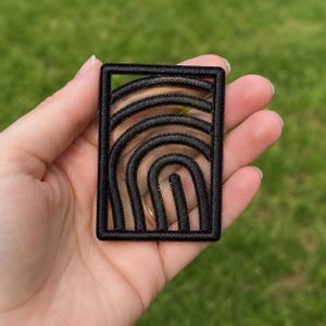 Pode incluir: Um emblema retangular bordado em preto apresentando uma série de linhas arqueadas concêntricas dentro de uma borda sólida. O design consiste em costuras pretas elevadas e texturizadas que criam um padrão de arco dentro da moldura.