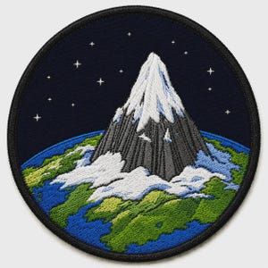 May include: A circular embroidered patch featuring a dark blue night sky with white stars above a snow-capped grey mountain peak. The mountain sits atop a stylized view of the Earth, showing green landmasses and blue oceans with white clouds.