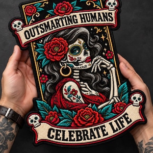 May include: A rectangular embroidered patch featuring a skeleton figure with sugar skull makeup, long dark hair, and red flowers. The design includes two banners with the text OUTSMARTING HUMANS and CELEBRATE LIFE, accented by small skulls and teal leaves.