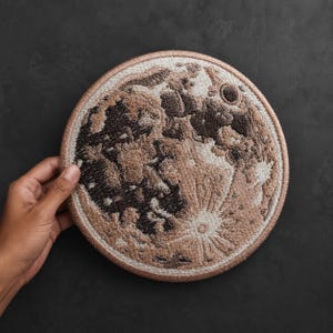 Puede incluir: Un parche bordado circular que presenta una representación detallada de la luna. El diseño utiliza hilos en tonos marrón, bronceado, crema y carbón oscuro para crear la superficie lunar, incluyendo cráteres y rayos, con un borde cosido.