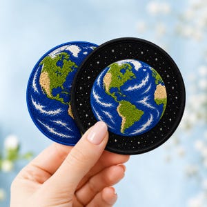 Puede incluir: Dos parches bordados circulares con una representación detallada de la Tierra. Un parche tiene un borde azul sólido, mientras que el otro presenta un borde negro con pequeñas estrellas blancas bordadas que rodean el planeta con tierras verdes y océanos azules.