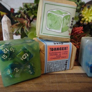 May include: A translucent green cube-shaped soap containing multi-sided dice with white numbers. Next to it is a wrapped soap bar with a label reading !DANGER! Gelatinous Cube Handle at Own Risk Contains Dice, and a blue translucent soap cube.