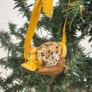 May include: A decorative ornament featuring a small yellow sculpted figure resting inside a walnut shell. The figure has a brown fabric shell with a white floral pattern on its back. A yellow ribbon is attached for hanging the ornament.