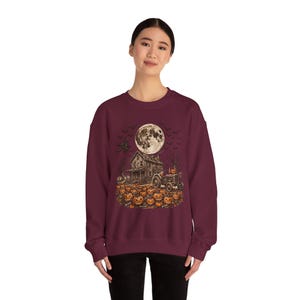 May include: A maroon crewneck sweatshirt featuring a graphic print of a spooky Halloween scene with a full moon, a dilapidated house, a tractor, and a field of carved jack-o'-lantern pumpkins under a sky filled with silhouettes of flying bats.