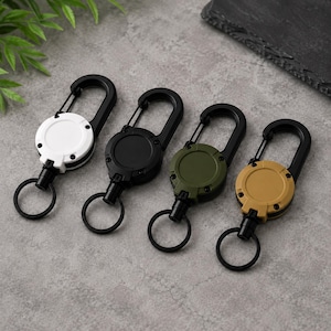 May include: Four retractable keychains with black carabiners, each featuring a circular housing in white, black, olive green, or tan. Each unit includes a small black split ring attached to a retractable cord, designed for securing keys or small tools.