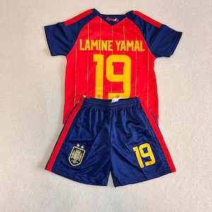 May include: A red and blue soccer jersey and shorts set featuring the name LAMINE YAMAL and the number 19 in yellow. The jersey has the word España near the neckline, and the shorts display the number 19 and a Spanish national team crest.