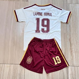 May include: A white soccer jersey with maroon trim and the text LAMINE YAMAL and the number 19 in maroon on the back. Paired with maroon soccer shorts featuring yellow side stripes, the number 19 in yellow, and a Spanish national team crest.