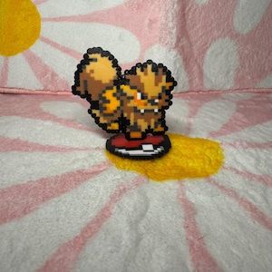 Può includere: Una figura in pixel art della creatura Arcanine realizzata con piccole perline di plastica fuse. La figura presenta colori arancione, nero e crema e poggia su una base circolare progettata per assomigliare a una Poké Ball rossa e bianca.