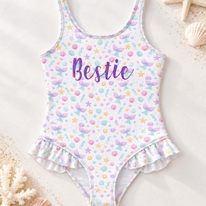 Op de afbeelding: Een wit badpak uit één stuk met een herhalend patroon van paarse zeemeerminstaarten, roze schelpen, gele zeesterren en lichtblauwe zeepaardjes. Het woord Bestie is in paars schrift op de borst gedrukt en de beenopeningen zijn voorzien van ruches.